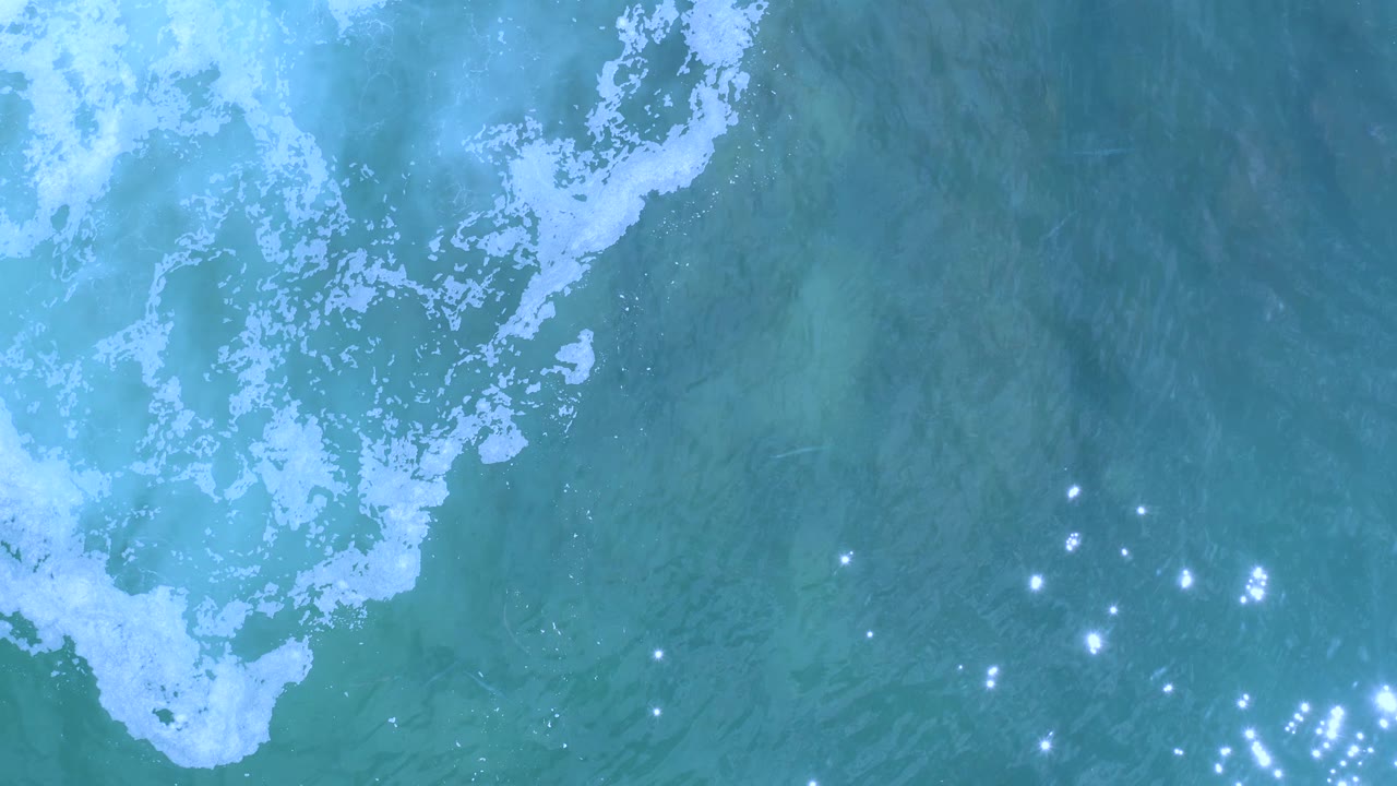 Overhead Los Cabos water frame with a broad white foam band near the top and a cluster of sun sparkles lower in the clear seawater, no visible people.