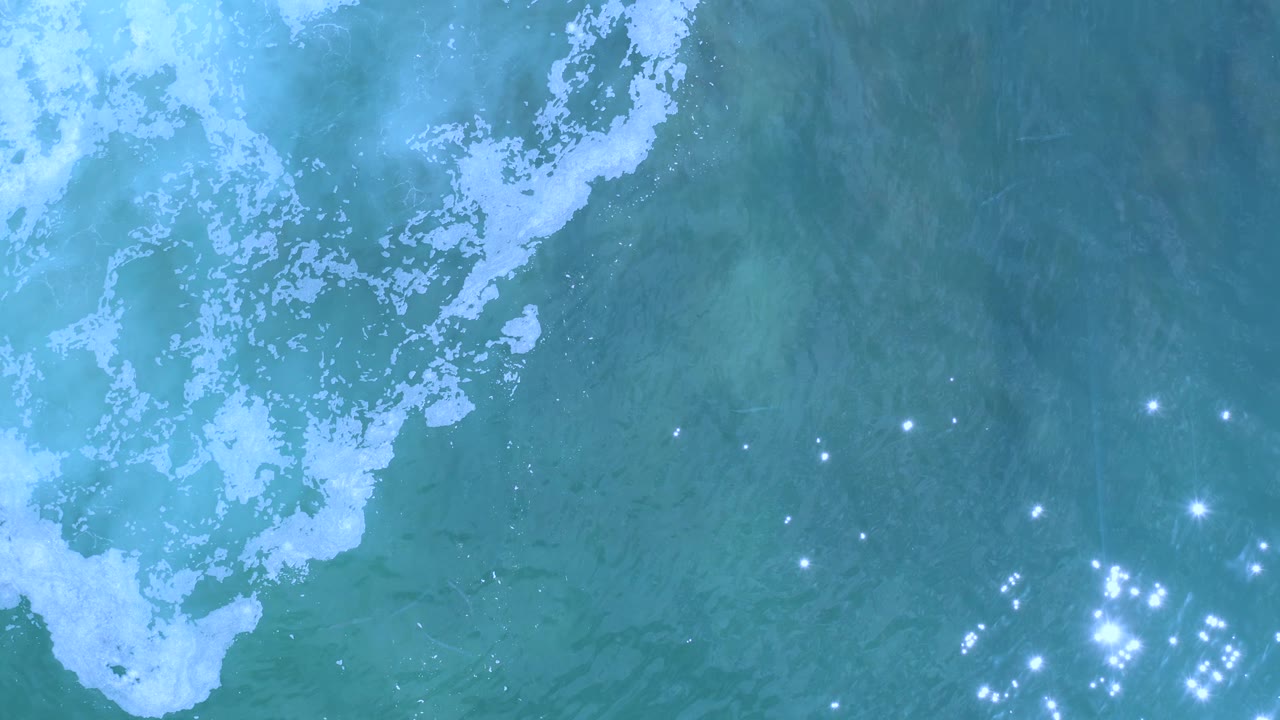 Top-down view of blue-green Los Cabos seawater with white foam along the upper edge and bright sun glints gathered toward the lower right, no visible people.