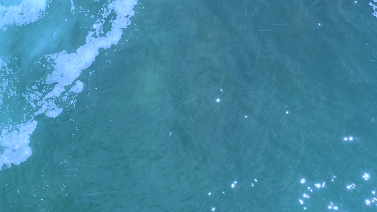 High view of blue-green ocean water in Los Cabos, Mexico, with a white foam patch near the upper left and small sun glints across the surface, no visible people.