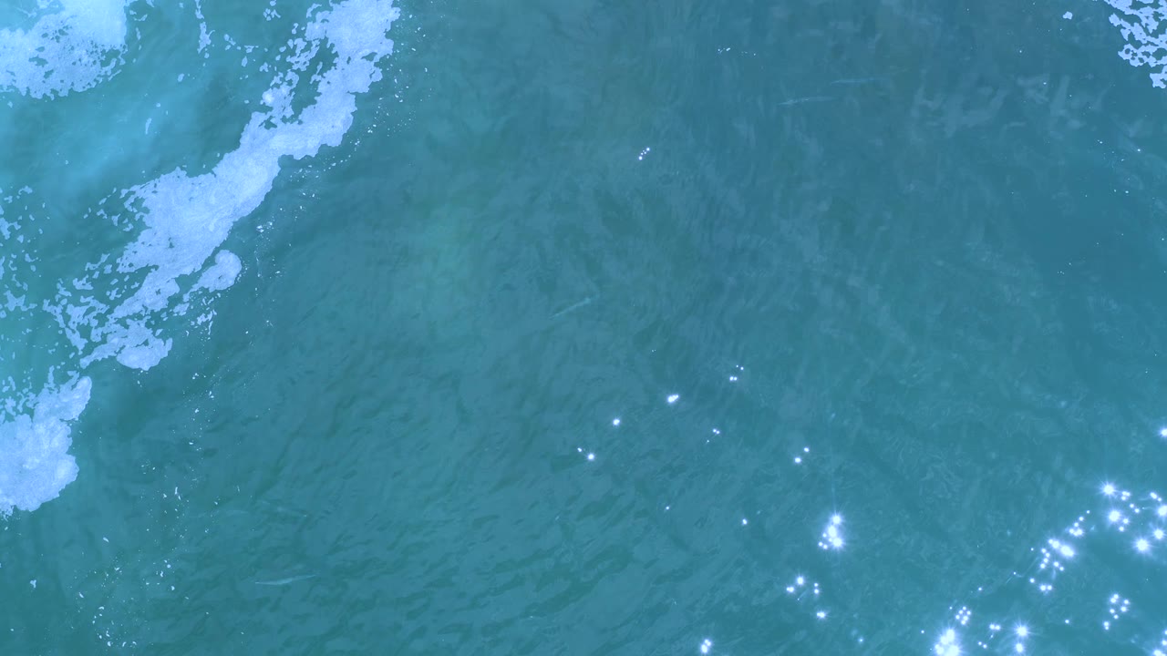 Overhead view of Los Cabos ocean water with a narrow foam patch near the top and bright sparkles beginning to cluster toward the lower right, no visible people.