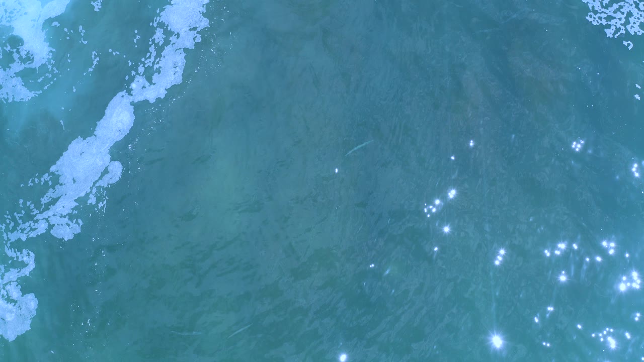 Overhead view of blue-green ocean water in Los Cabos, Mexico, with a white foam streak along the left side, scattered sun glints near the lower center, and no visible people.