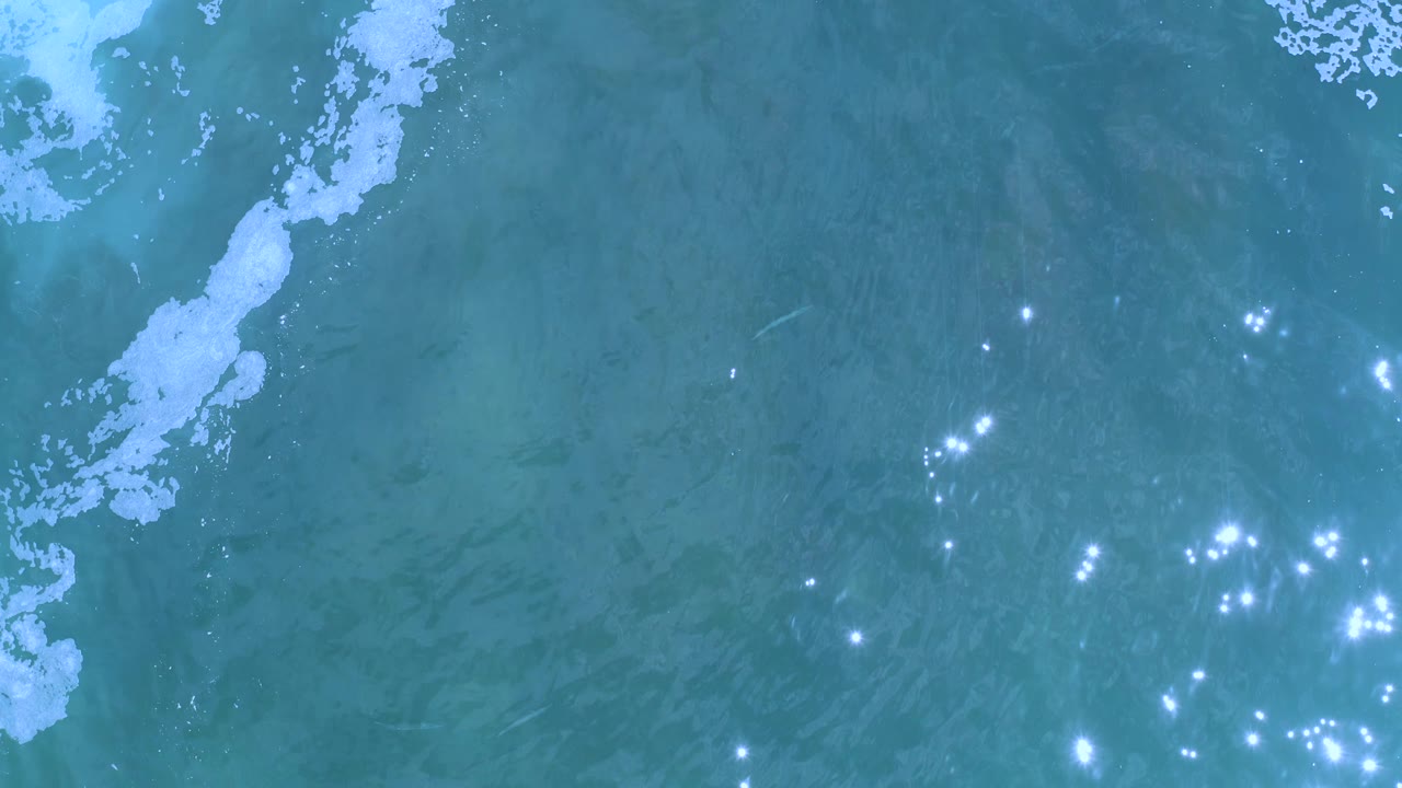 Overhead Los Cabos ocean frame with a pale foam band near the upper left and a tighter cluster of bright reflections in the lower half, with no visible people.