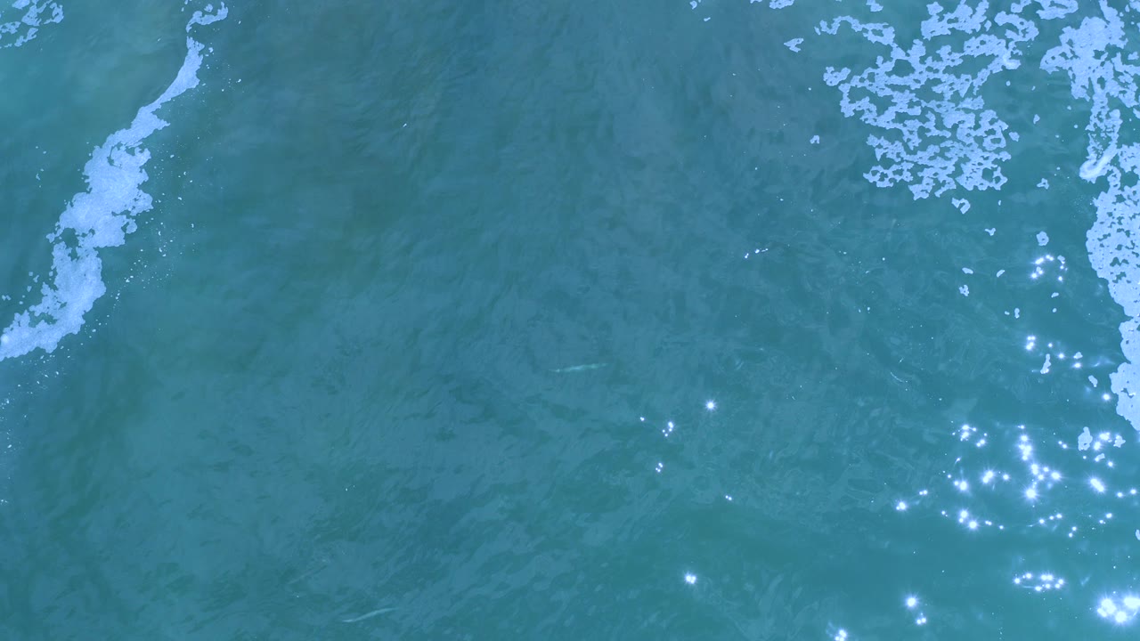 Overhead view of blue-green ocean water in Los Cabos with white foam entering from the top edge, scattered sun reflections near the lower center, and no visible people.