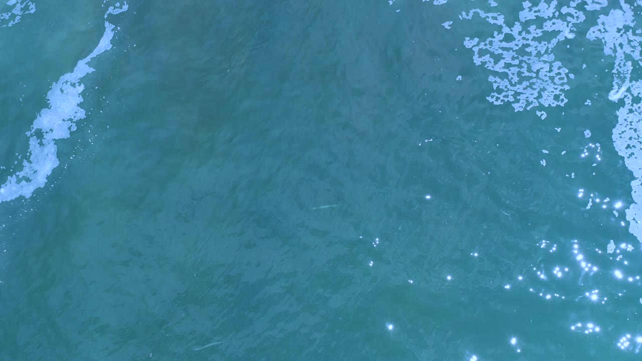Aerial Los Cabos ocean frame with pale foam clustered along the upper edge and small bright sun glints scattered across the lower half, with no visible people.