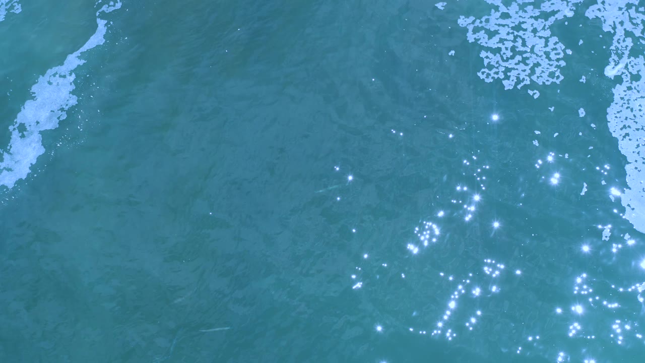 Top-down Los Cabos water view with turquoise surface texture, foam patches near the upper edge and right side, and dense sun sparkle across the lower right, with no visible people.