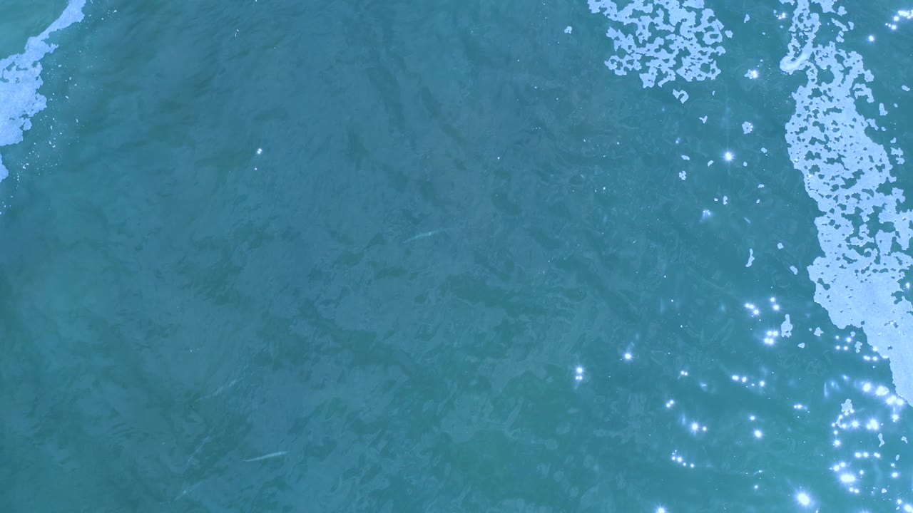 Los Cabos ocean surface seen from above, with a tall band of white foam dropping from the upper center-right and small sun glints across the lower water, with no visible people.