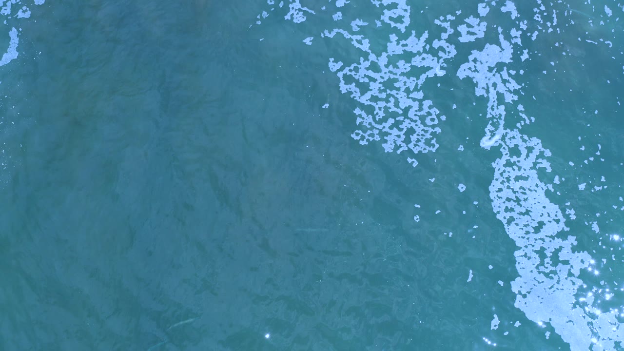 Overhead teal ocean water with a vertical white foam trail.