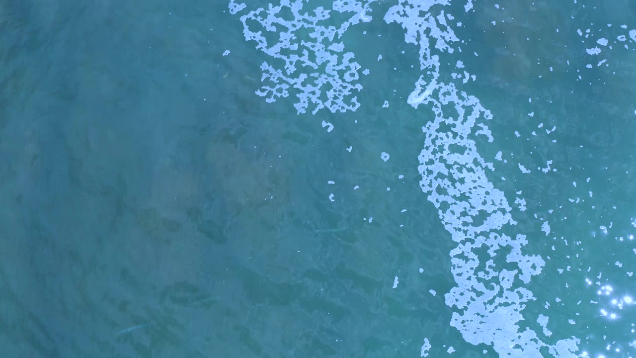 Overhead Los Cabos ocean frame with the white bubble trail running down the center-right of the blue-green water and bright reflections near the bottom edge, no visible people.