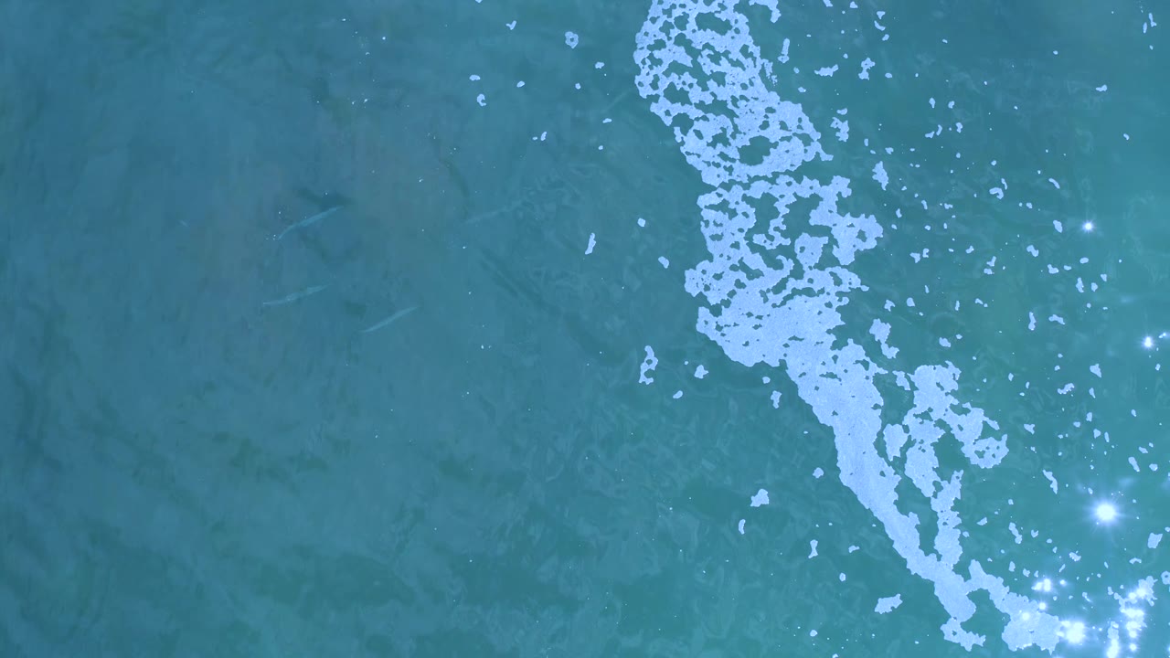 Overhead Los Cabos water surface with white foam running diagonally through the frame and clustered light reflections near the lower right, with no visible people.