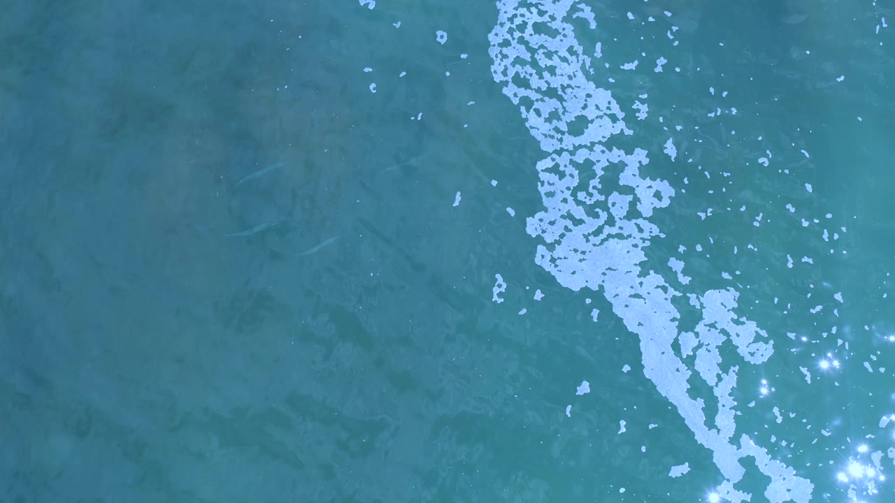 Overhead ocean texture in Los Cabos, Mexico, with the white foam band shifted toward the upper right and scattered sun glints across the water, with no visible people.