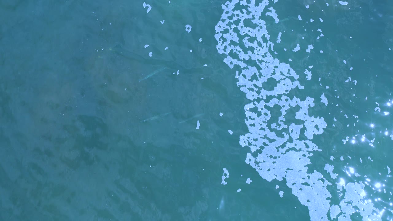 Overhead view of teal ocean water in Los Cabos, Mexico, with a pale foam streak near the center-right, small sun sparkles, and no visible people.