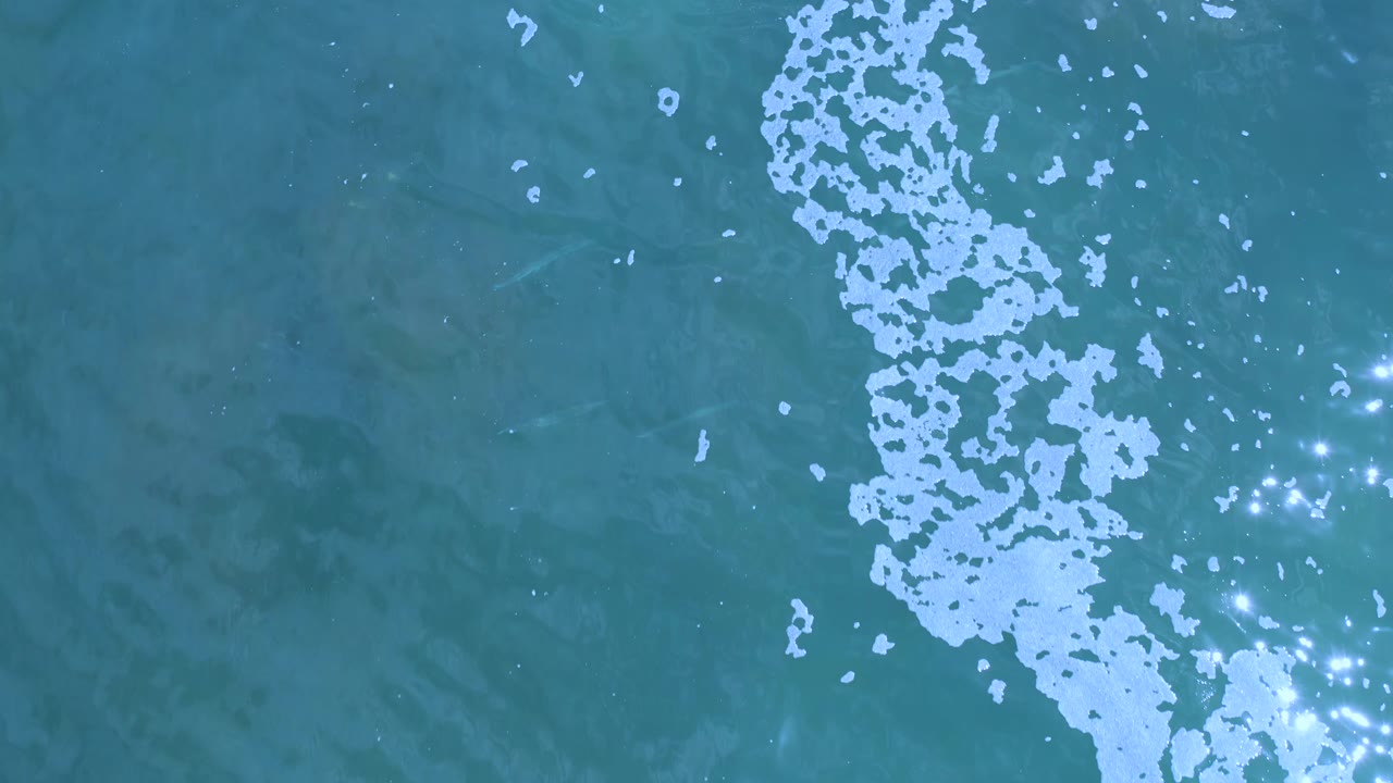 Blue-green Los Cabos ocean water fills the frame, with the white foam band shifted slightly through the middle and bright glints clustered to the right, no visible people.