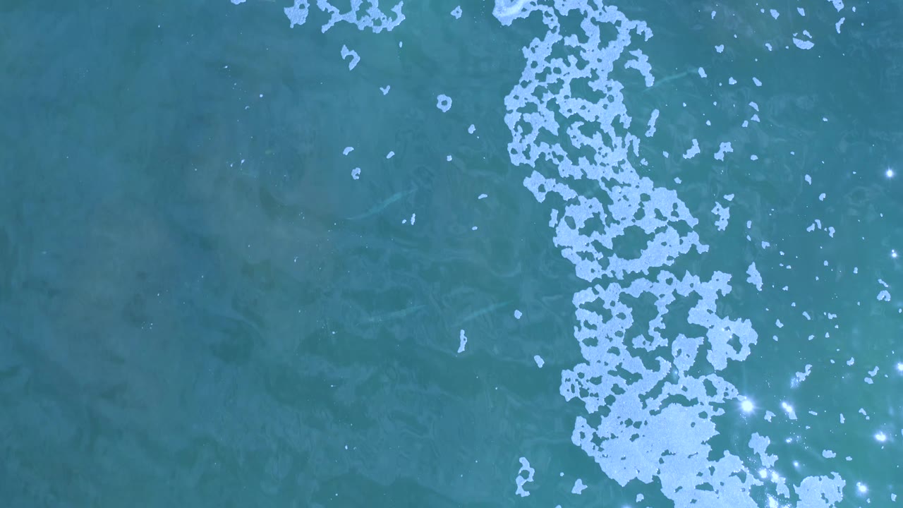 Aerial view of teal water in Los Cabos, Mexico, where pale foam runs down the right side and scattered white specks sit over darker green water, no visible people.