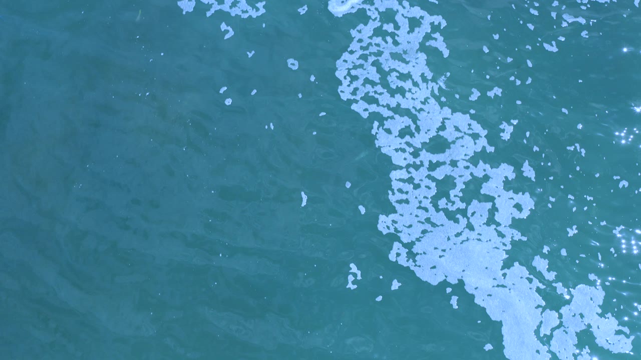 Overhead view of turquoise water in Los Cabos, Mexico, with a vertical band of white foam near the center and small sun reflections on the surface, with no visible people.