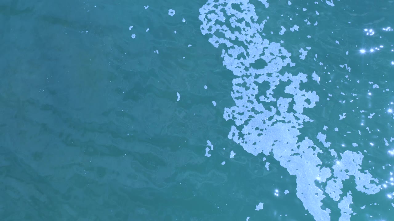 Straight-down view of teal water in Los Cabos, Mexico, with an irregular white foam line entering from the upper right and scattered sun flashes, with no visible people.