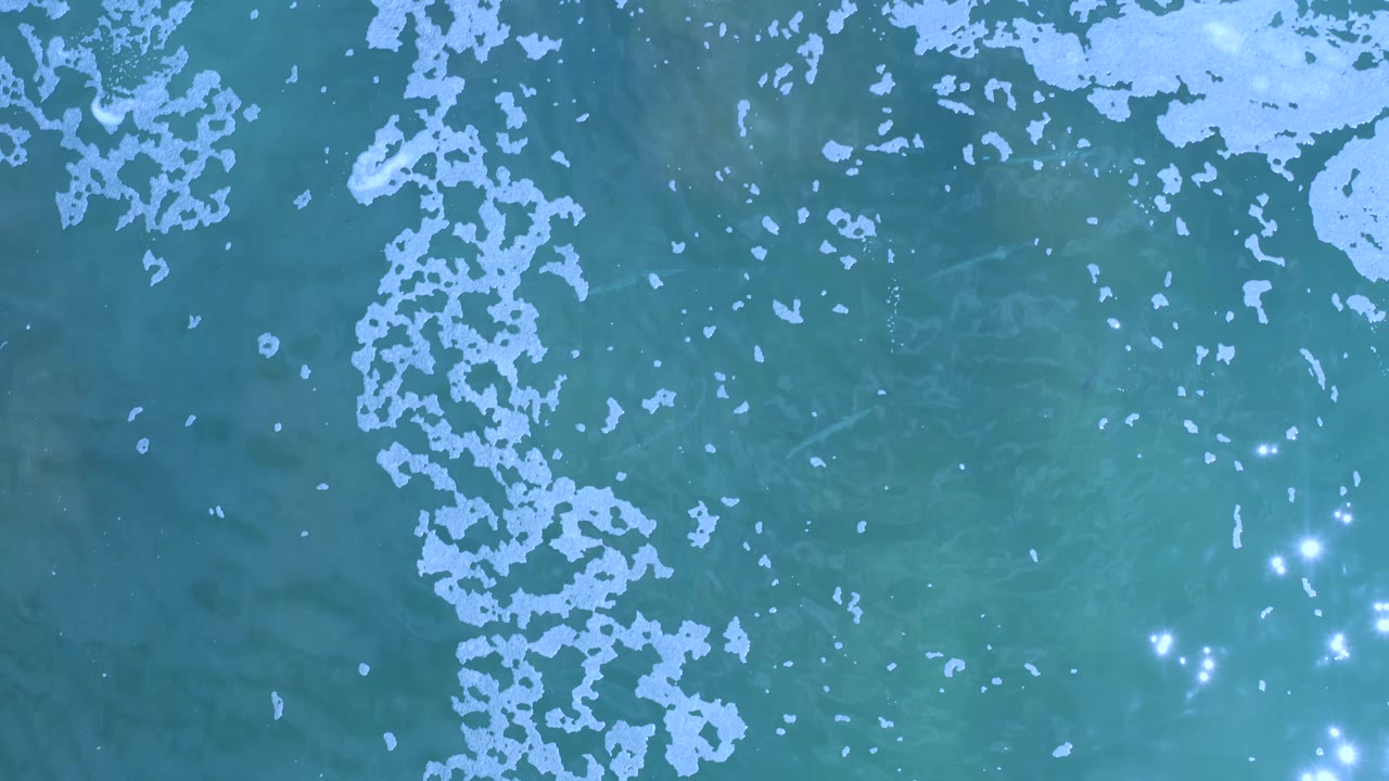 Aerial Los Cabos water surface with a vertical band of pale foam near the left side, small sun reflections to the right, and no visible people.