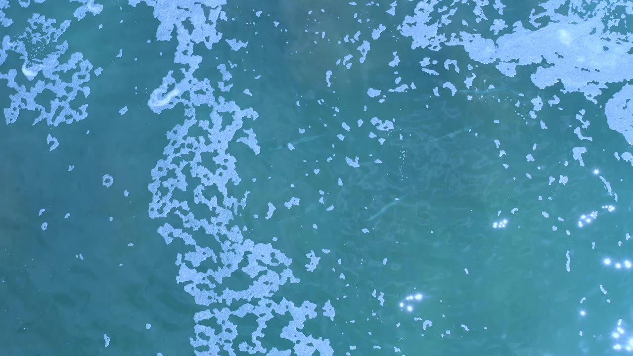 Overhead ocean frame from Los Cabos with scattered white foam across the top and left, teal water below, bright glints at right, and no visible people.