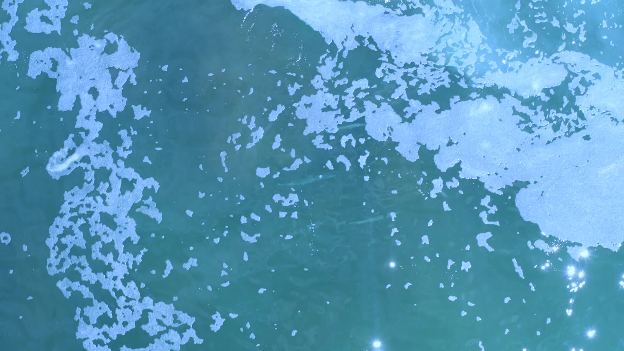 Overhead view of blue-green sea in Los Cabos with broken white foam near the top and left edges, scattered sun reflections on the water, and no visible people.