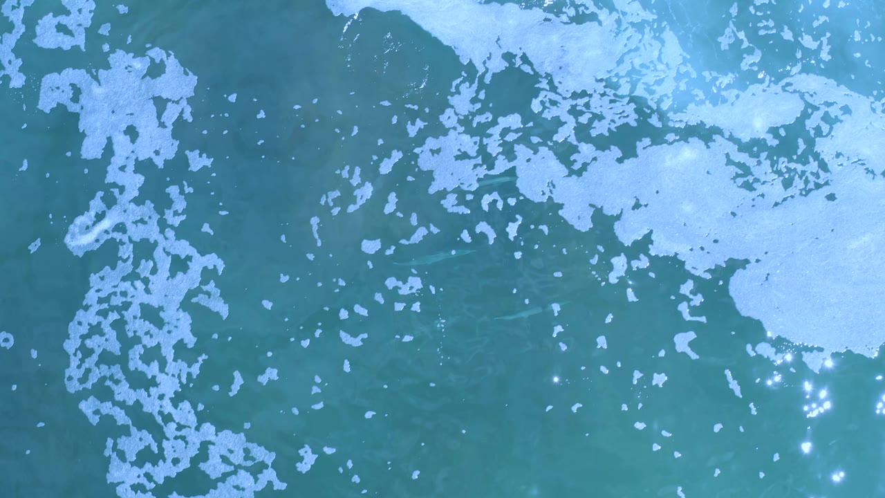Top-down Los Cabos sea surface with a larger pale foam area across the upper right, darker blue-green water below, small white foam flecks, and no visible people.