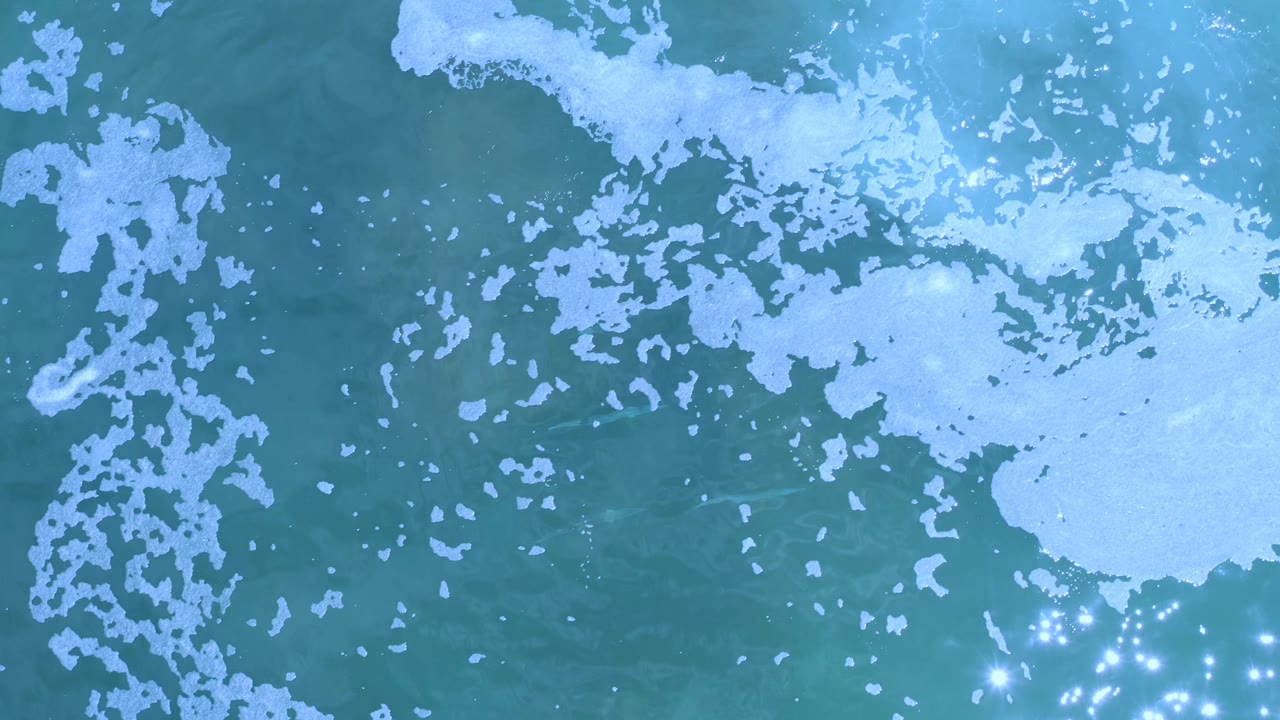 Aerial view over Los Cabos water with white foam clustered along the top and left side, turquoise sea through the center, bright sparkles near the lower right, and no visible people.