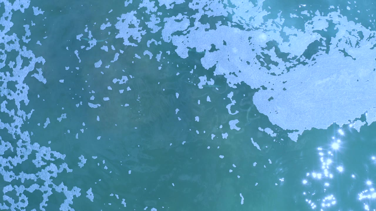Overhead view of blue-green sea in Los Cabos, Mexico, with scattered white foam near the upper frame and bright sun glints on the water, no visible people.
