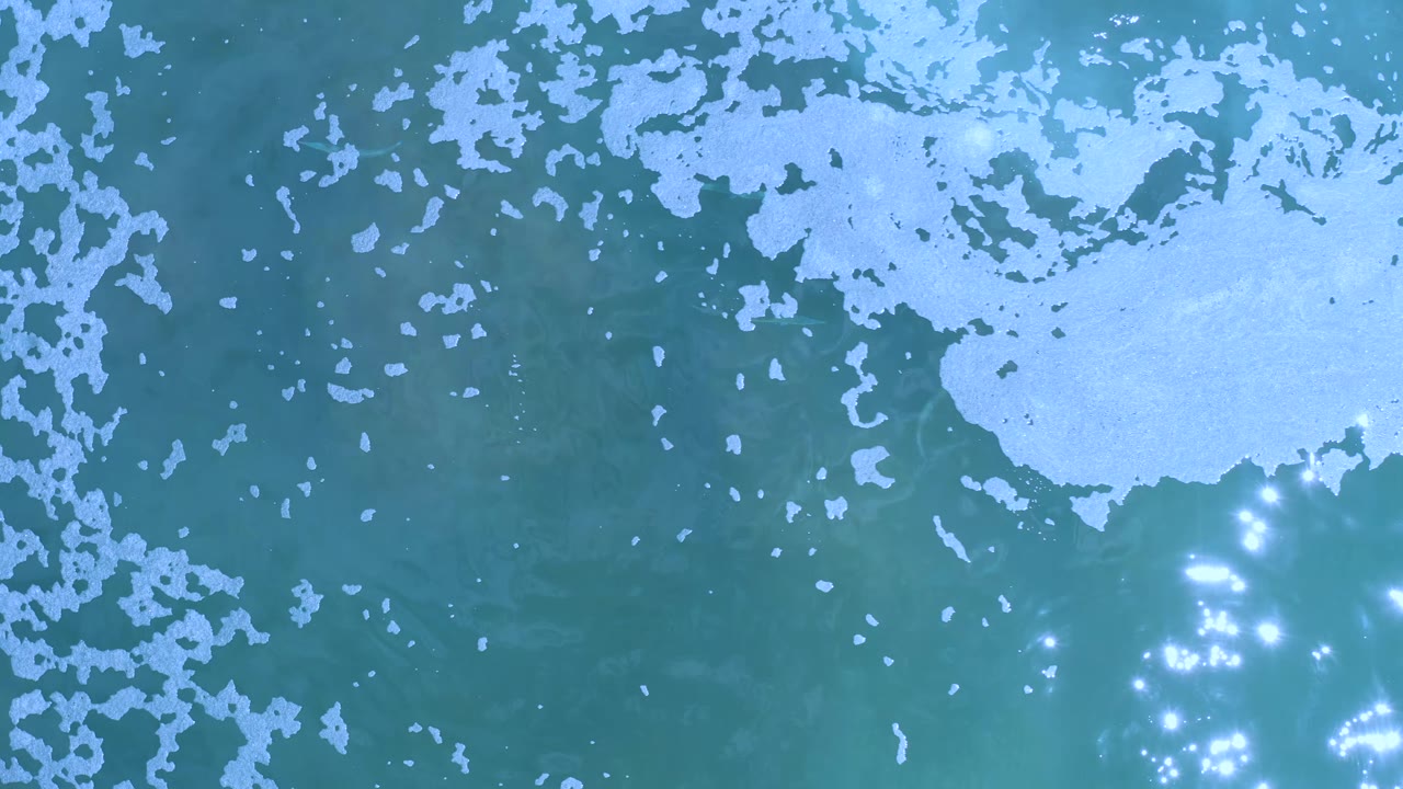 Blue-green sea surface in Los Cabos, Mexico, seen from above as white foam gathers across the upper and right areas with clustered light reflections, no visible people.