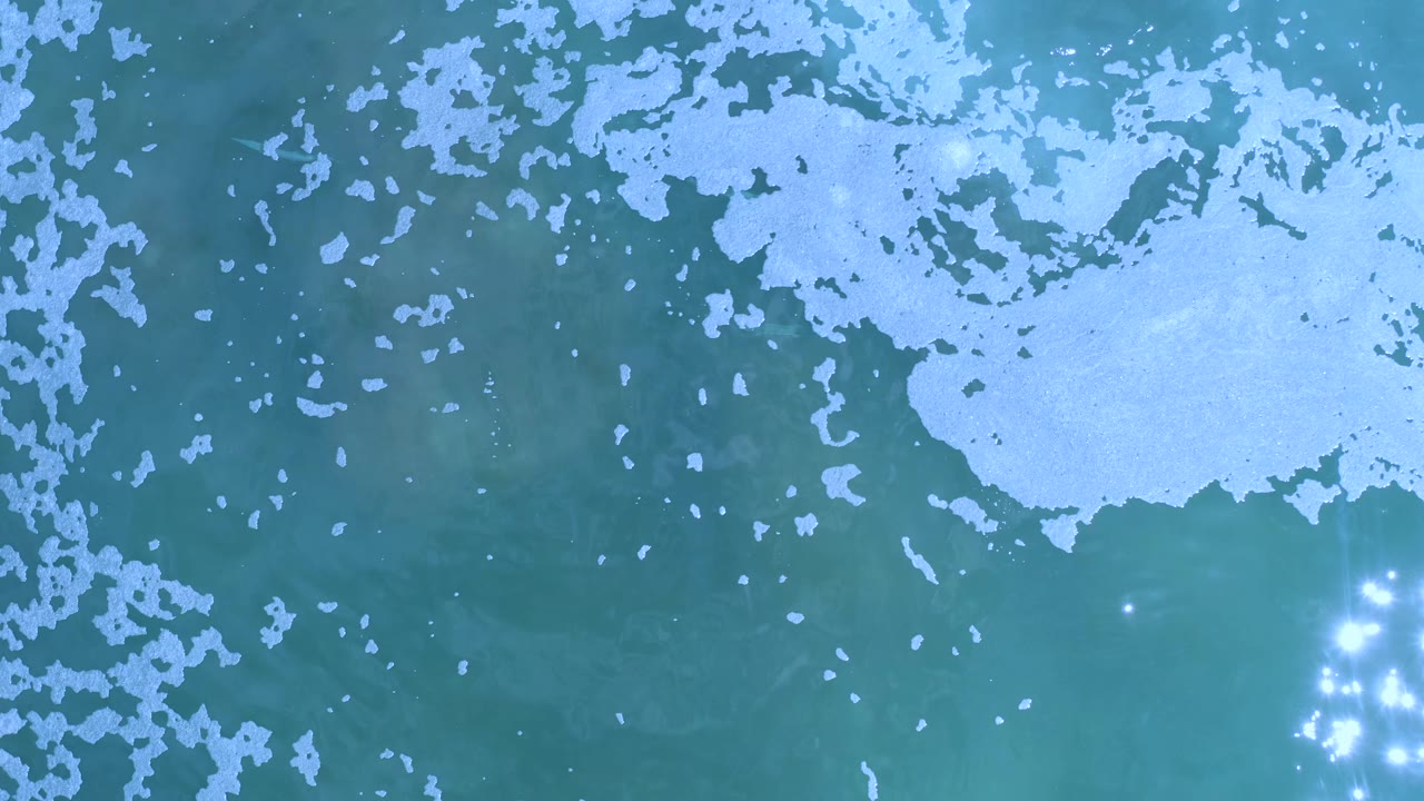 Overhead Los Cabos, Mexico sea view with pale foam along the top edge, darker blue-green water below, and a tight group of sun sparkles near the right side, no visible people.