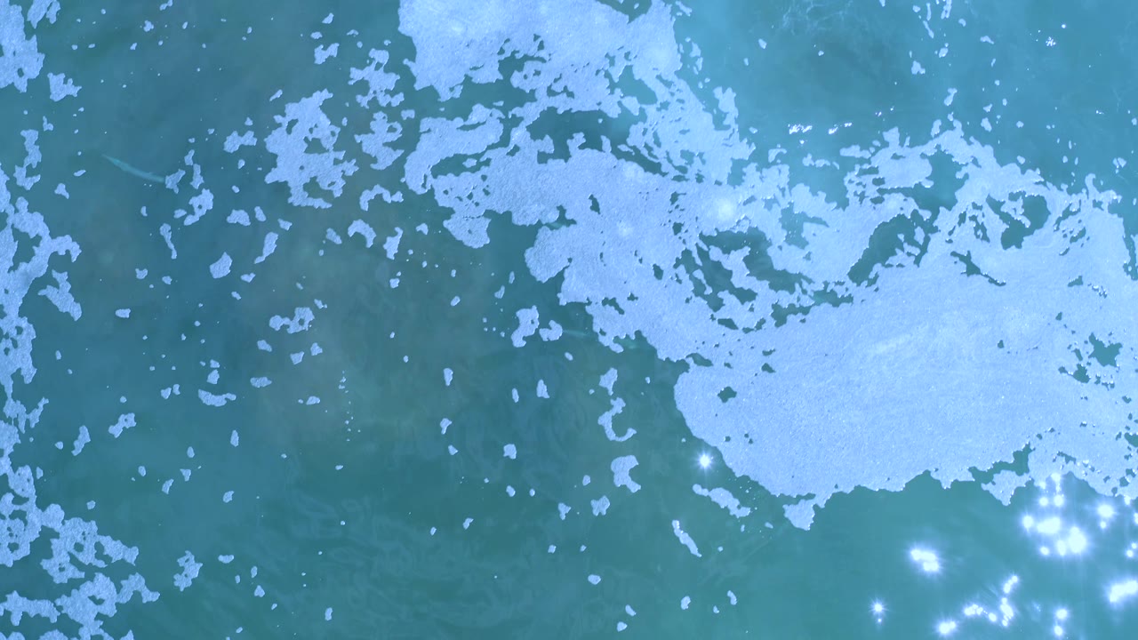 Aerial-looking view of Los Cabos, Mexico water with a broad white foam patch crossing the upper half and smaller flecks scattered through blue-green sea, no visible people.