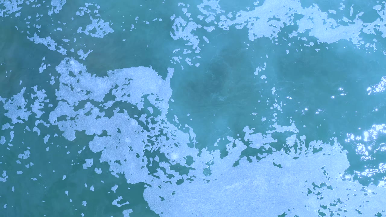 Overhead view of Los Cabos water with pale foam concentrated along the lower right edge and scattered bright sun reflections, with no visible people.