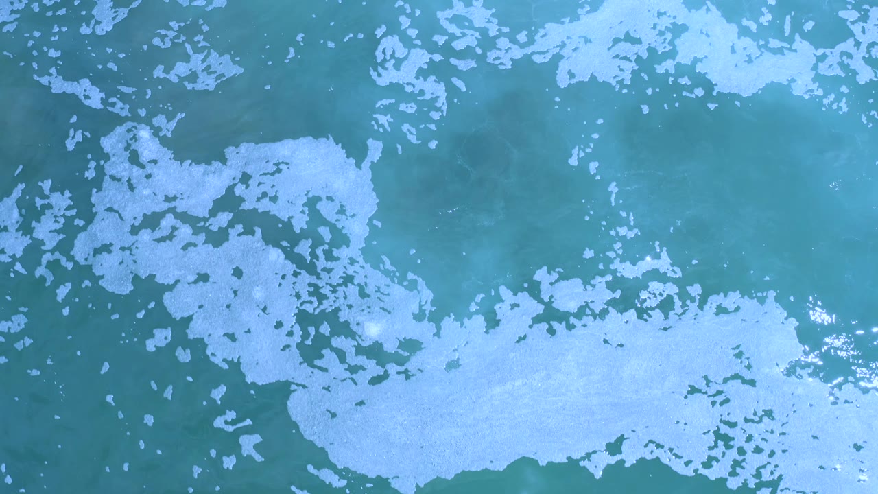 Overhead Los Cabos sea surface with a broad white foam patch along the lower portion and darker blue-green water above, with no visible people.