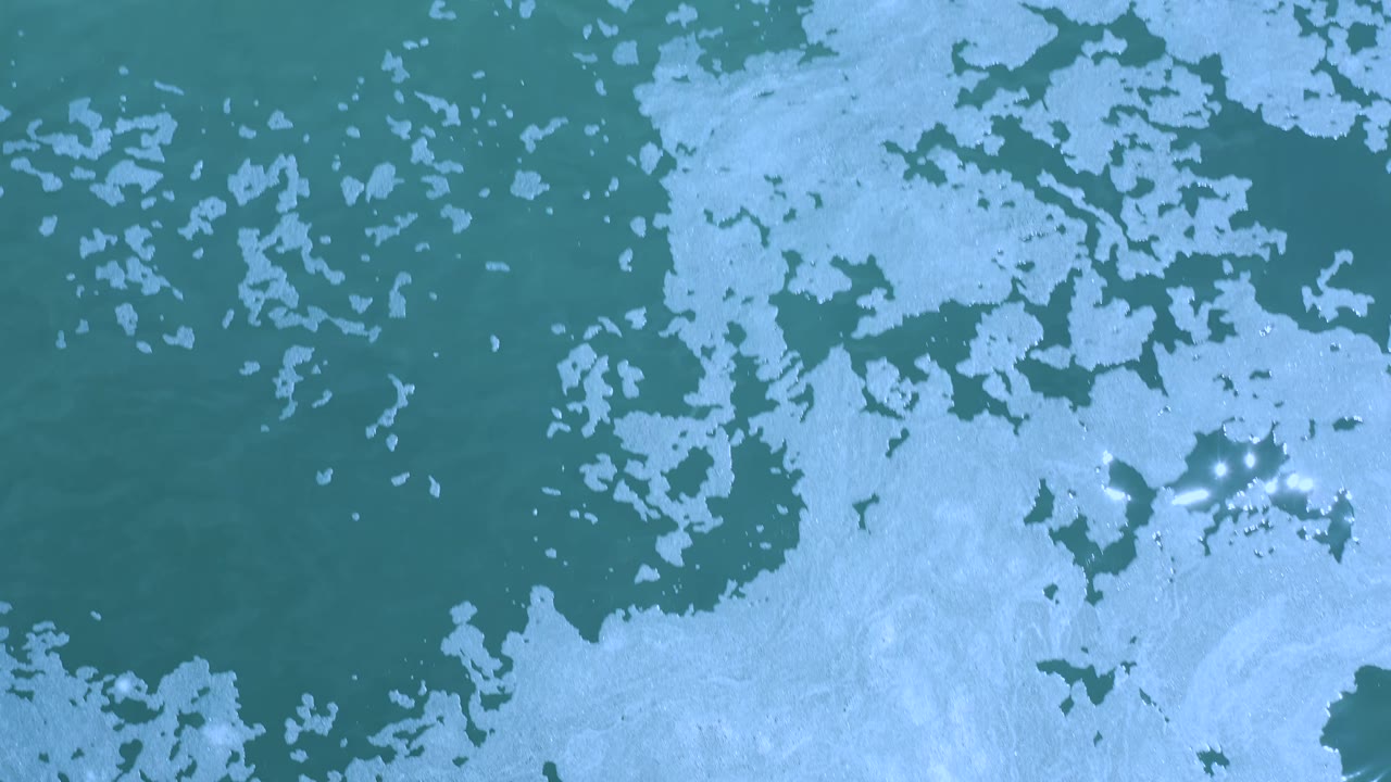 Aerial view of blue-green Los Cabos water with irregular foam shapes and several bright sun glints near the right side, with no visible people.