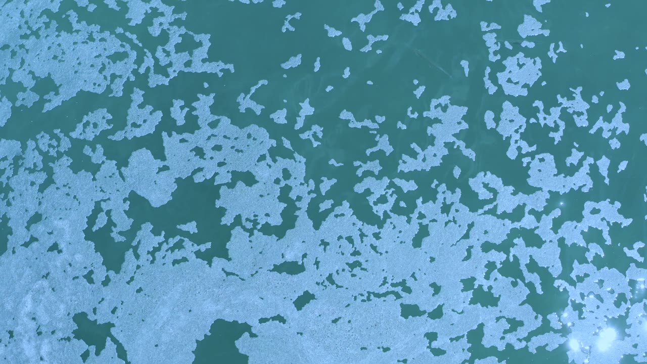White particles across blue-green water with a darker patch near the lower left.