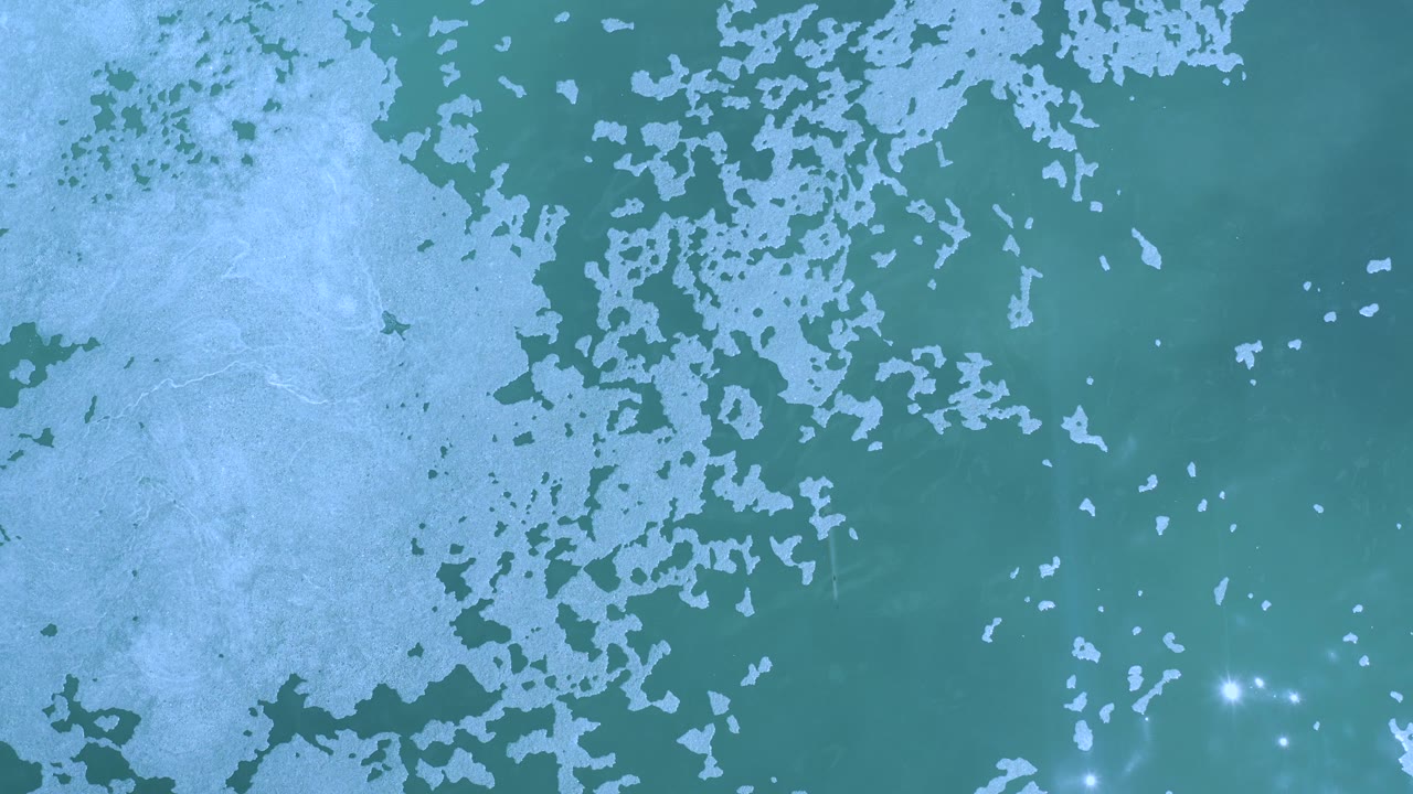 Opening overhead frame of turquoise sea in Los Cabos, Mexico with pale seafoam spreading across the left side, scattered sun glints near the lower edge, and no visible people.