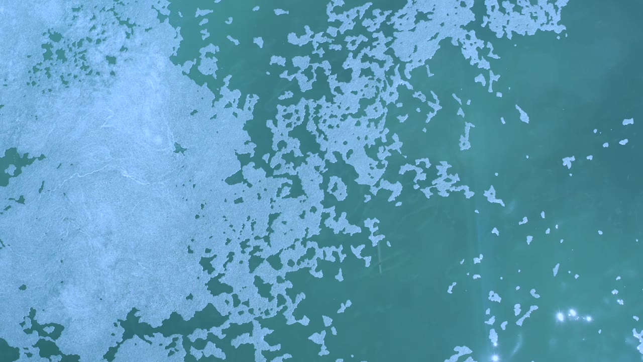 Top-down Los Cabos ocean frame with teal water, broken pale foam patches across the upper left and center, a few bright reflections near the bottom, and no visible people.