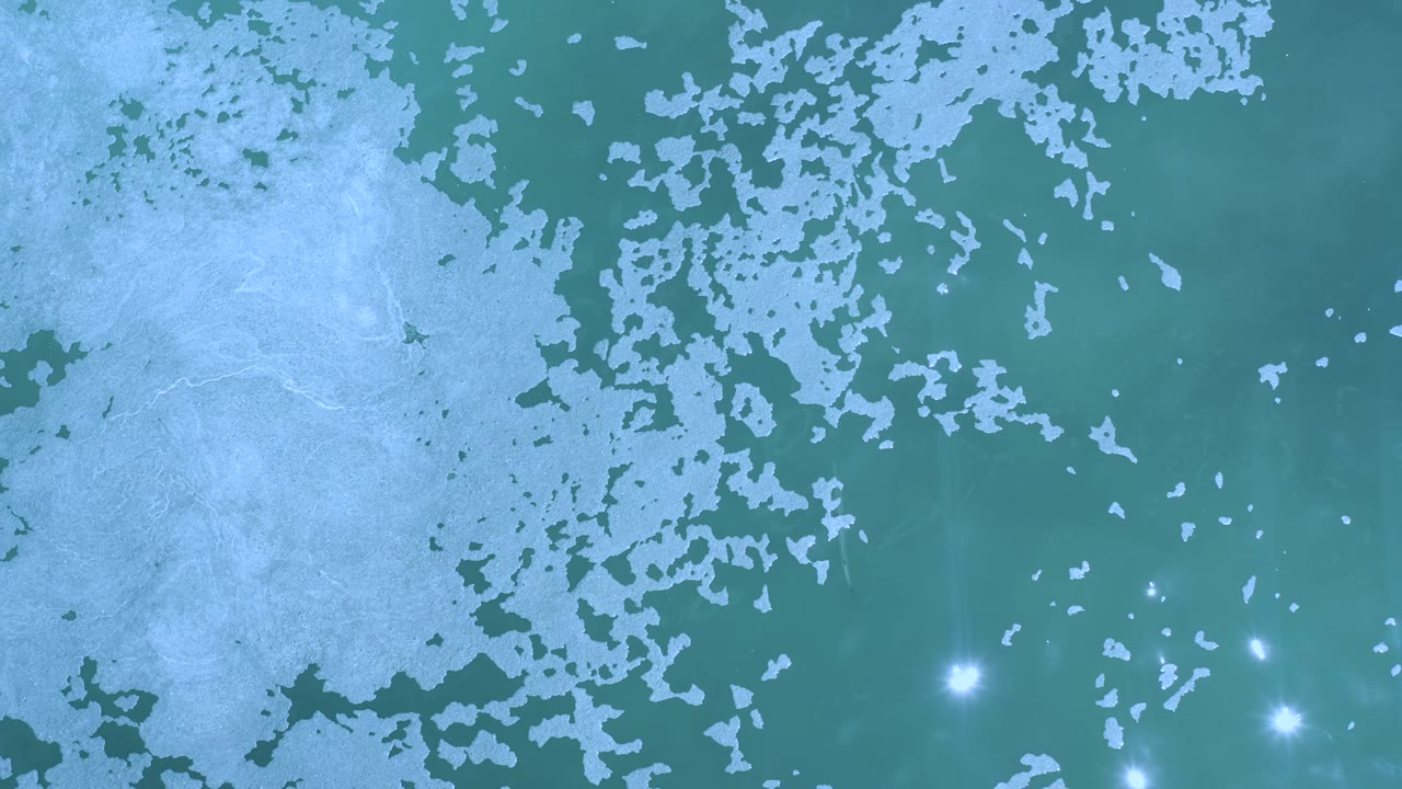 Aerial-looking Los Cabos water frame with green-blue sea, pale foam clustered across the upper half, isolated white glints toward the lower right, and no visible people.