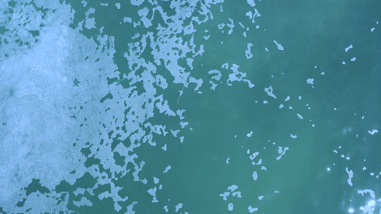 Overhead view of pale sea foam spreading across teal ocean water in Los Cabos, Mexico, with bright reflection spots near the lower edge and no visible people.