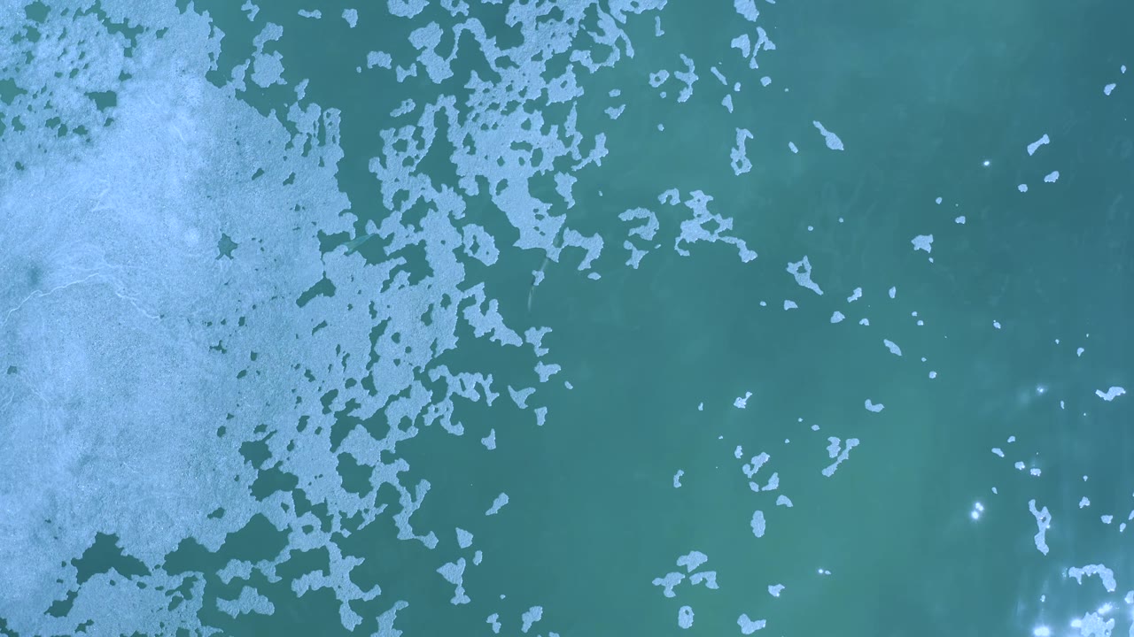 Top-down Los Cabos ocean frame with broken white foam patches across teal water and a loose cluster of small glare points near the bottom center, with no visible people.
