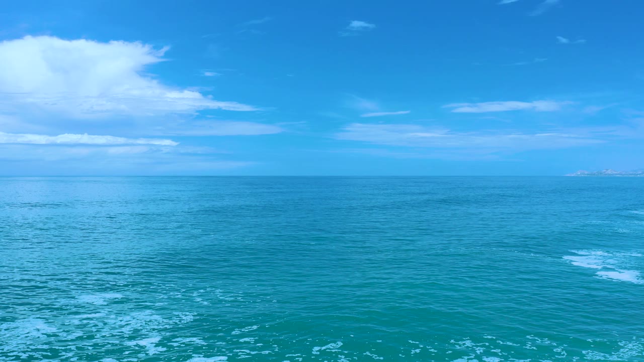 Los Cabos coastal water at 0:02, showing turquoise surface texture, a small patch of white foam at the right edge, distant land, and no visible people.