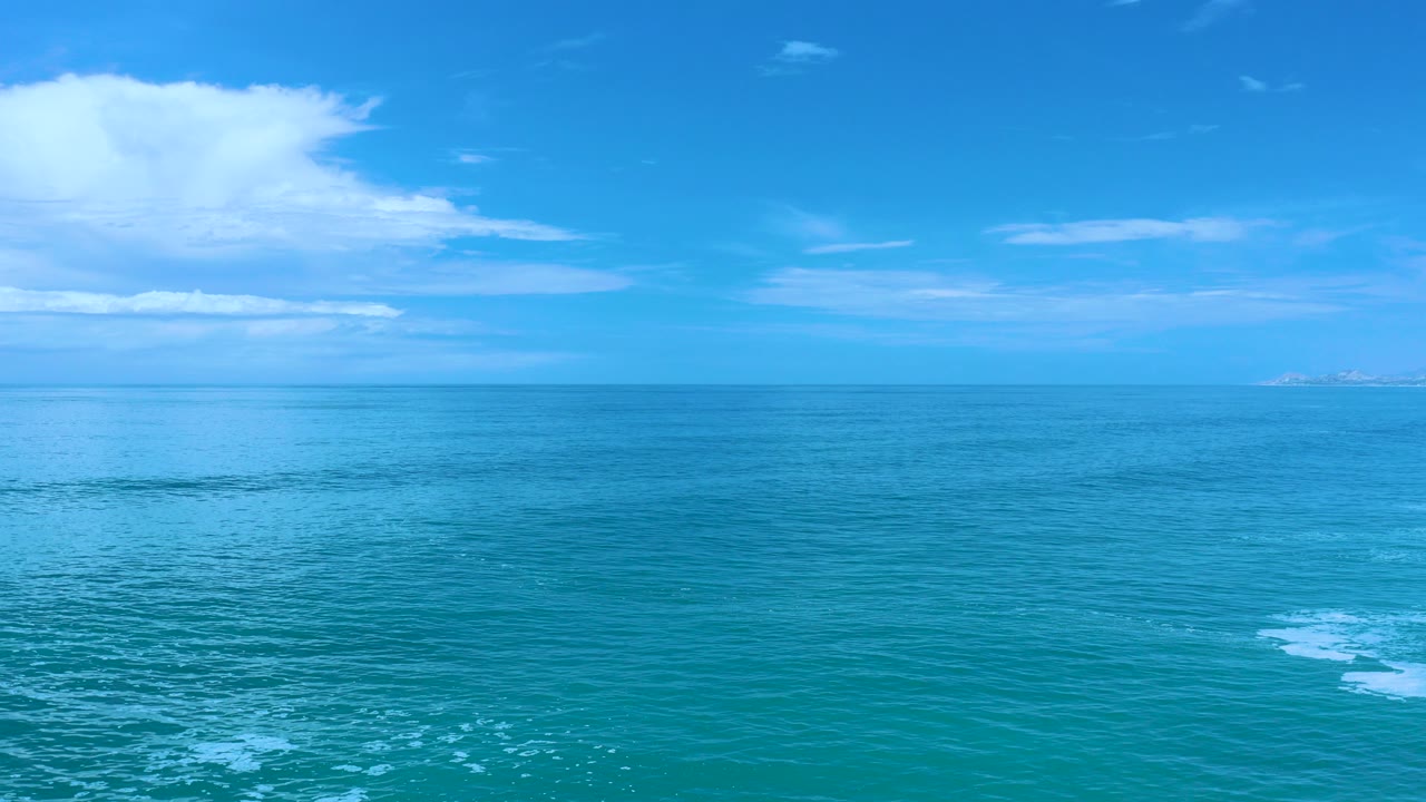 Wide ocean view in Los Cabos, Mexico, with turquoise water, a level horizon, scattered white clouds, faint distant land, and no visible people.