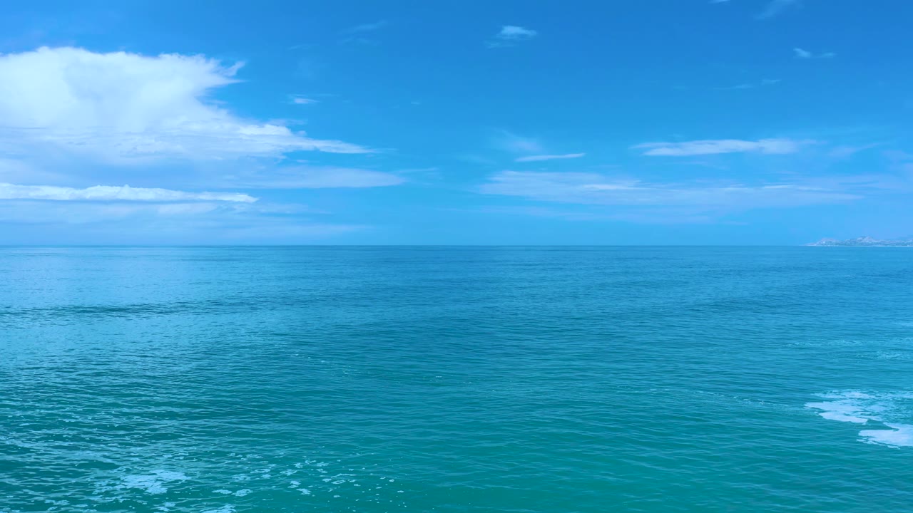 Los Cabos ocean frame with broad blue-green water, a soft cloud band overhead, faint land near the horizon, and no visible people.