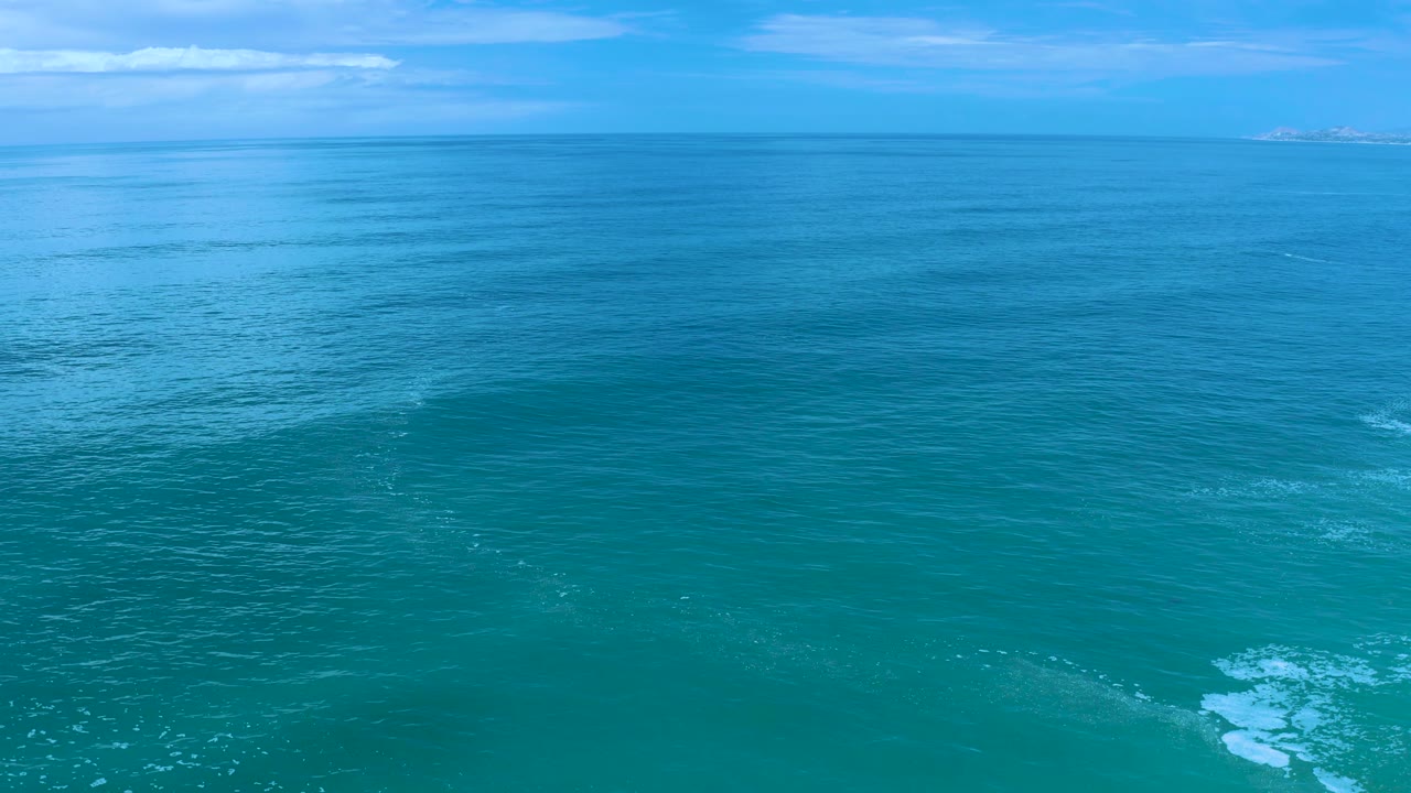 Wide sea view in Los Cabos, Mexico, with small white wave patches entering the lower frame, a pale blue sky, and no visible people.