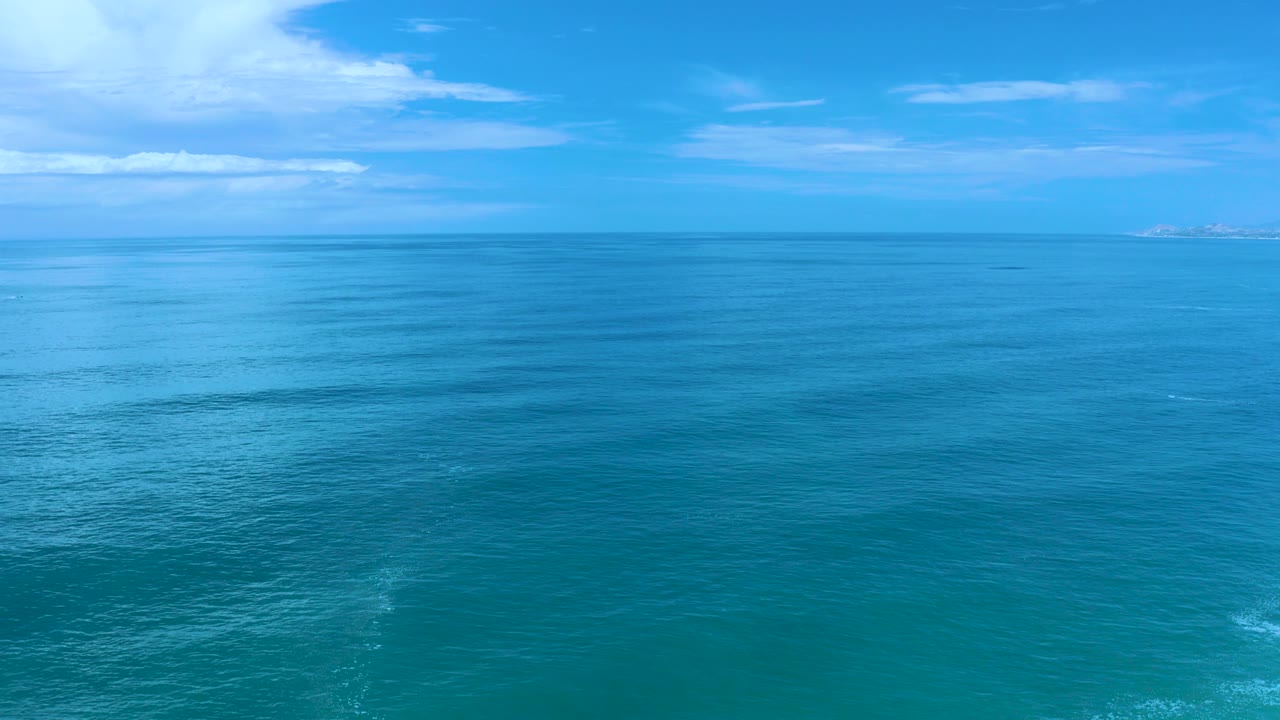 Wide Los Cabos ocean view at the start of the clip, with blue water under a bright sky, white clouds at left, faint land on the horizon, and no visible people.