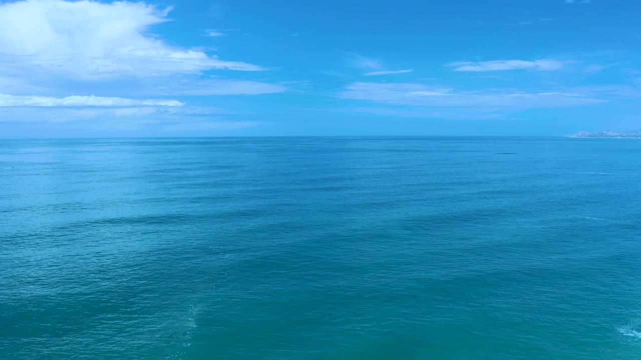 Mid-clip Los Cabos view with the ocean stretching to a level horizon, small white clouds overhead, faint land at the far right, and no visible people.