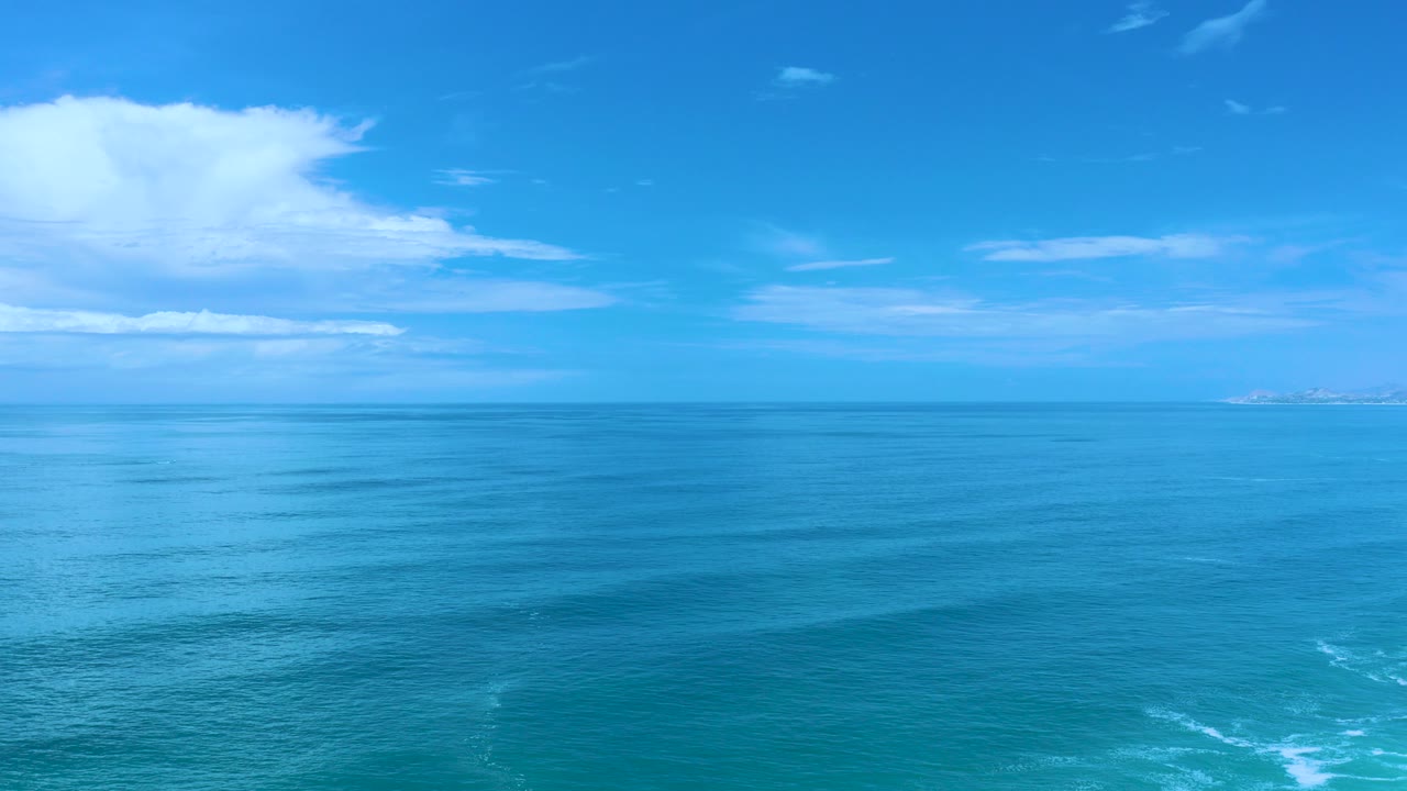 Opening frame of Los Cabos blue ocean with a level horizon, distant land at the far right, scattered white cloud, and no visible people.