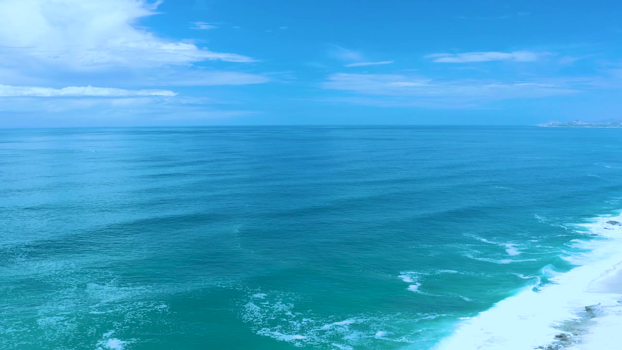 Los Cabos, Mexico ocean frame with a level horizon, layered blue water, white breakers reaching in from the lower right, thin clouds, and no visible people.