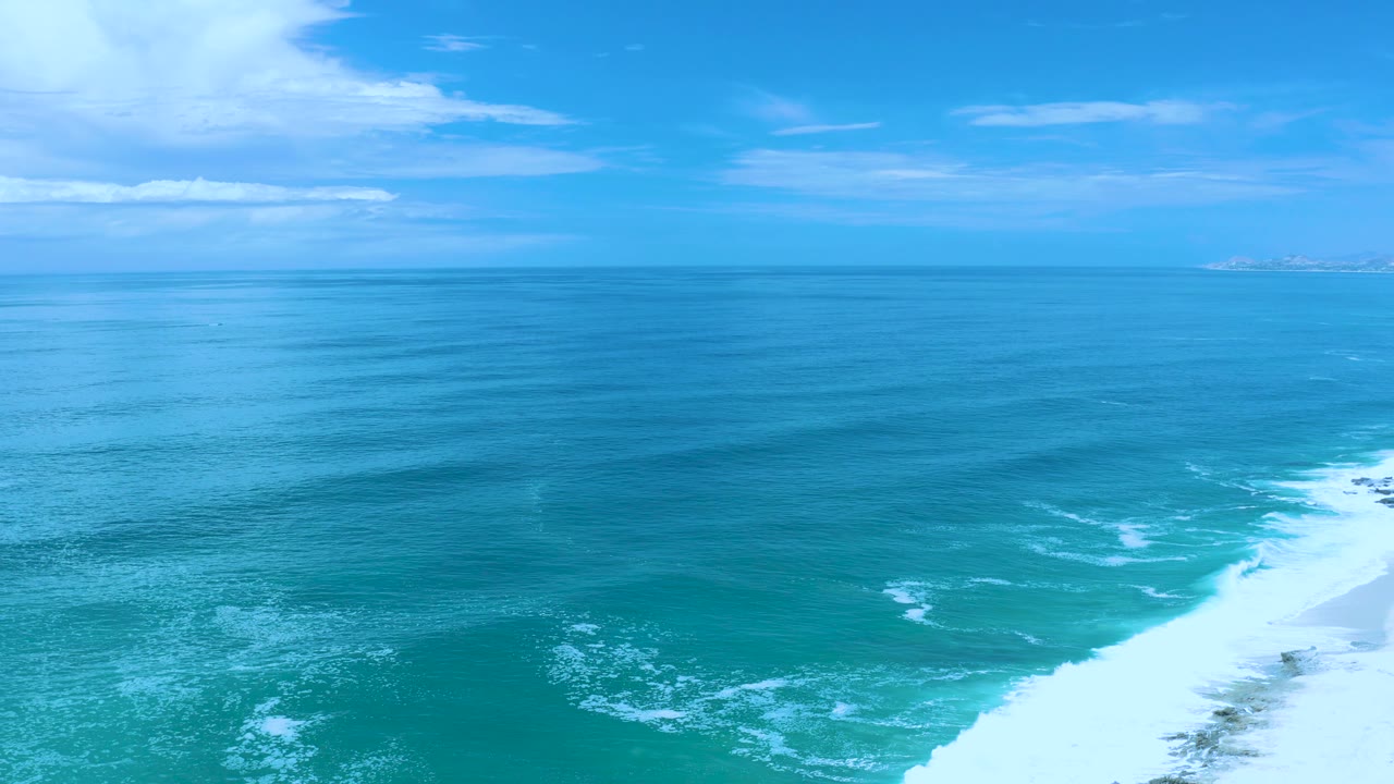 High coastal view in Los Cabos, Mexico, showing calm blue-green water, faint wave bands, a white surf line on the right, hazy distant clouds, and no visible people.