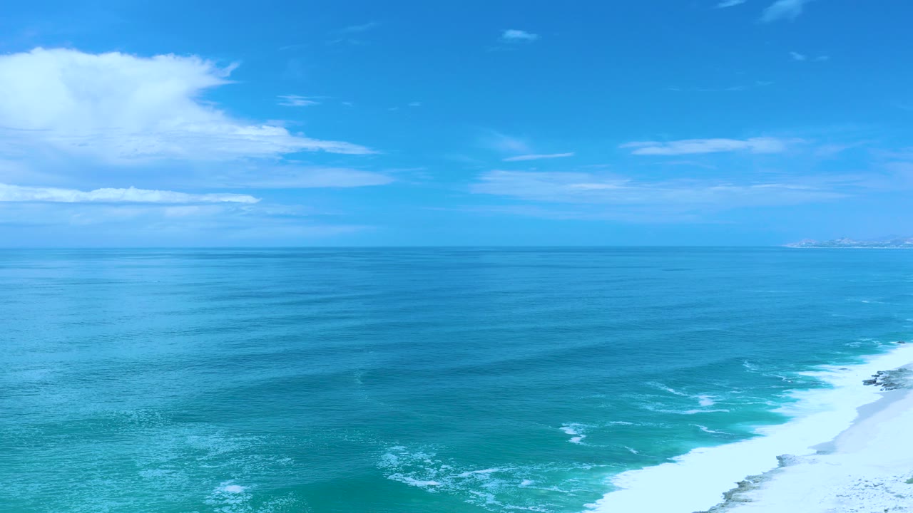 Wide aerial view of blue ocean, sky, surf, and beach edge