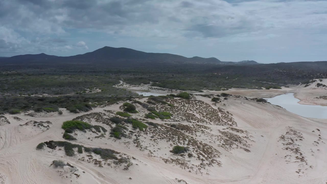 Wide aerial landscape with dunes, green scrub, water pools, and dark mountain ridge.