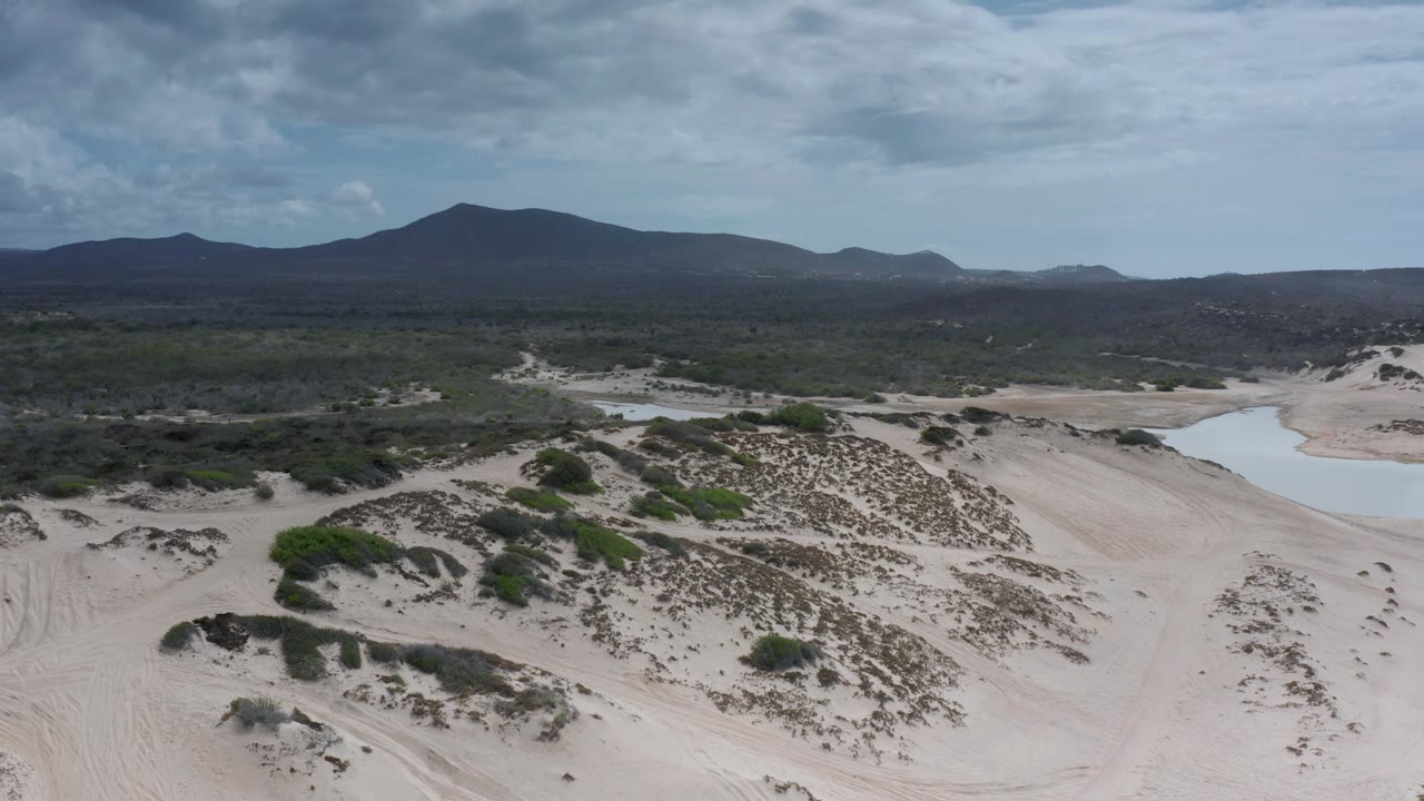 Los Cabos aerial frame with a shallow water pool beside pale sand dunes, dark green brush across the midground, distant mountains, cloudy sky, and no visible people.