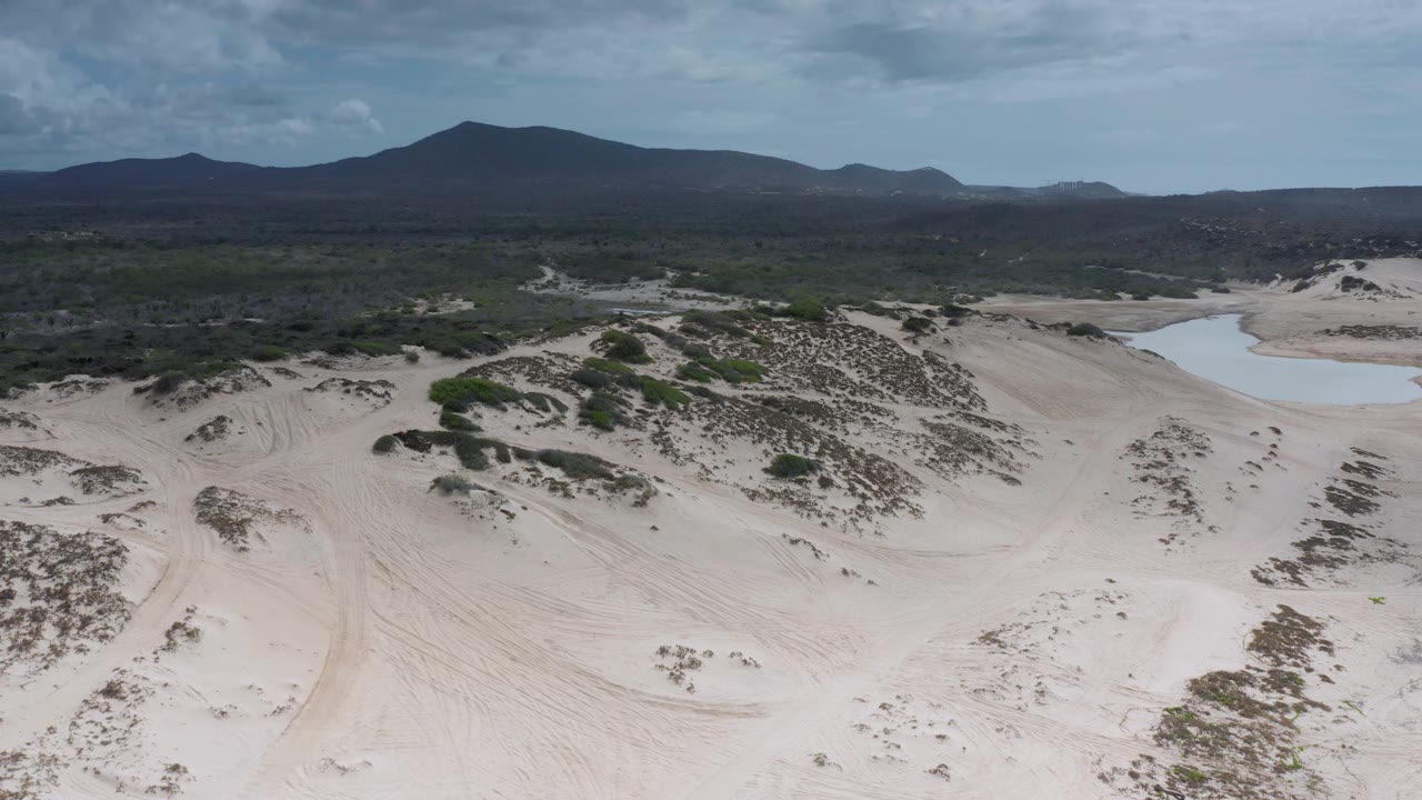Los Cabos aerial frame showing dune ridges and scrub patches across the center, the pond partly visible on the right, a broad dark mountain ridge on the horizon, and no visible people.